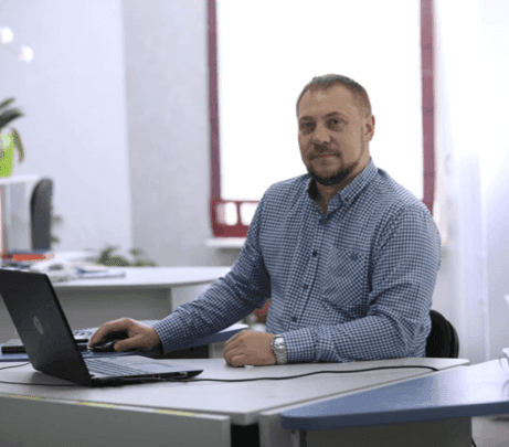 Man in a blue shirt sitting at a desk with a laptop, smiling at the camera.