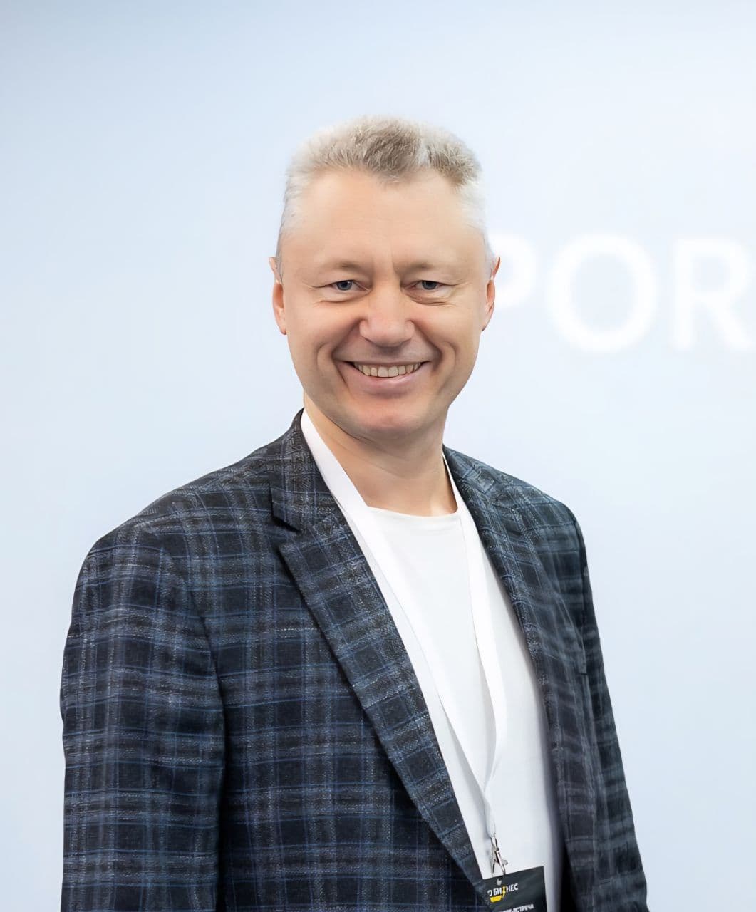 A smiling older man with gray hair and a plaid jacket looks directly at the camera.