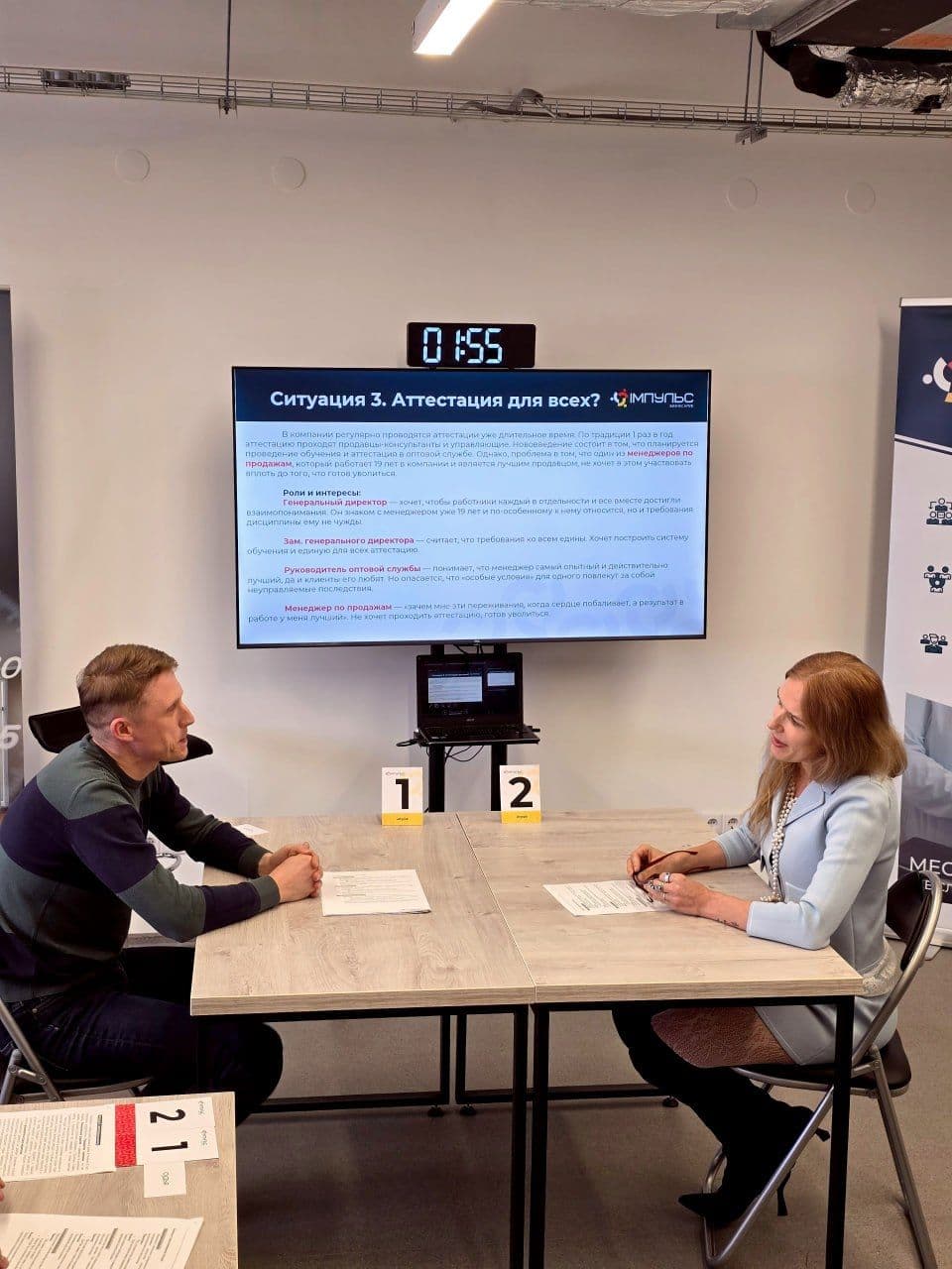 Two individuals sit across a table, facing a large screen displaying information.
