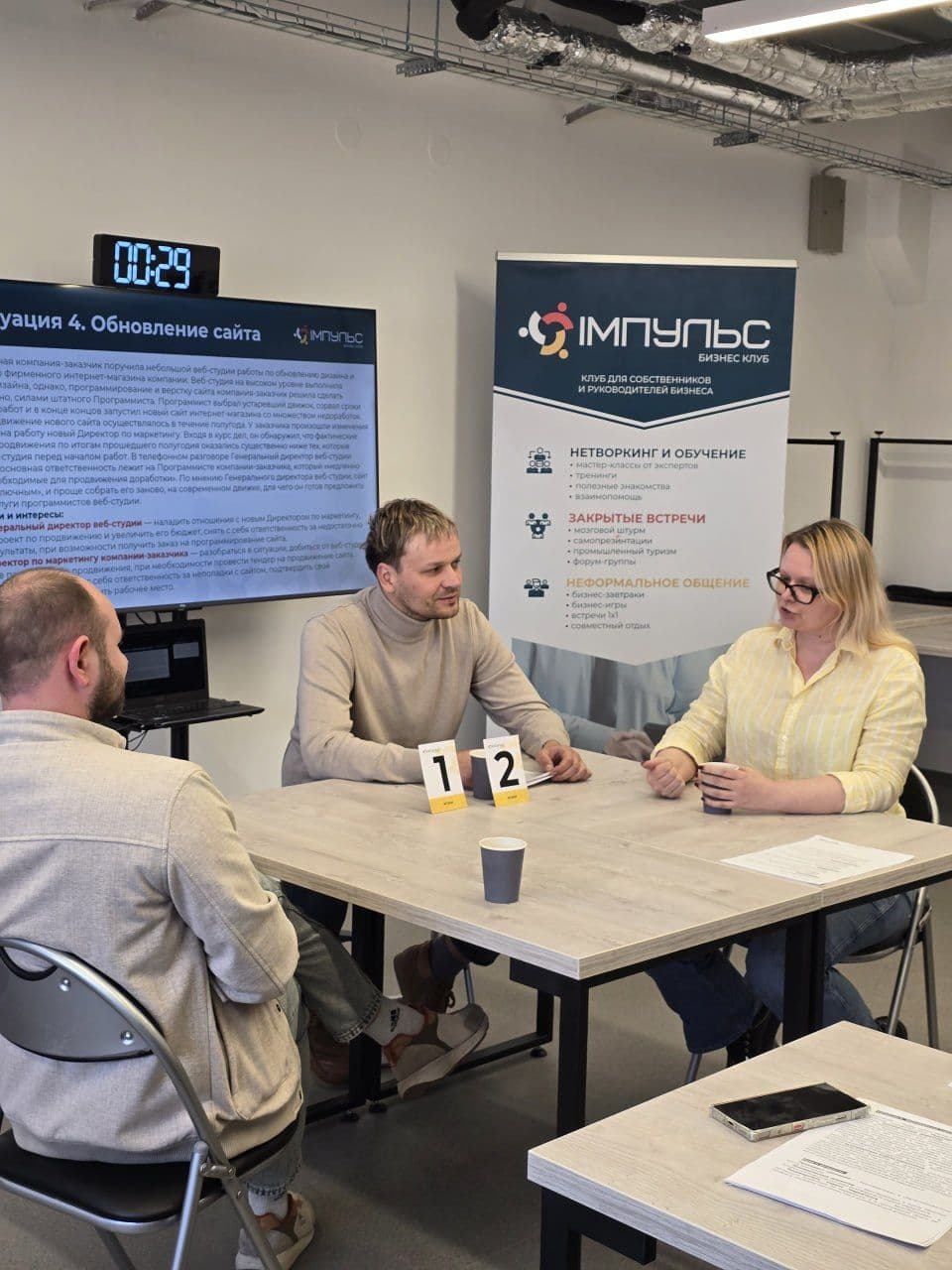 Three people engaged in conversation at a table with a screen and banner.